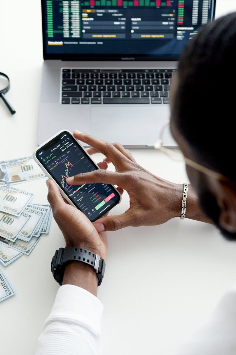 A person trading stocks using a smartphone, showcasing financial data on mobile and laptop screens.